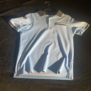 Blå pikétröja med kontrastdetaljer - Säljer en snygg ljusblå pikétröja med mörkare krage och ärmslut. Den har en knappslå framtill och en liten bröstficka med kontrastkant. Perfekt för en avslappnad men stilren look. Passar bra till både vardag och fest! Pris går alltid att förhandlas!