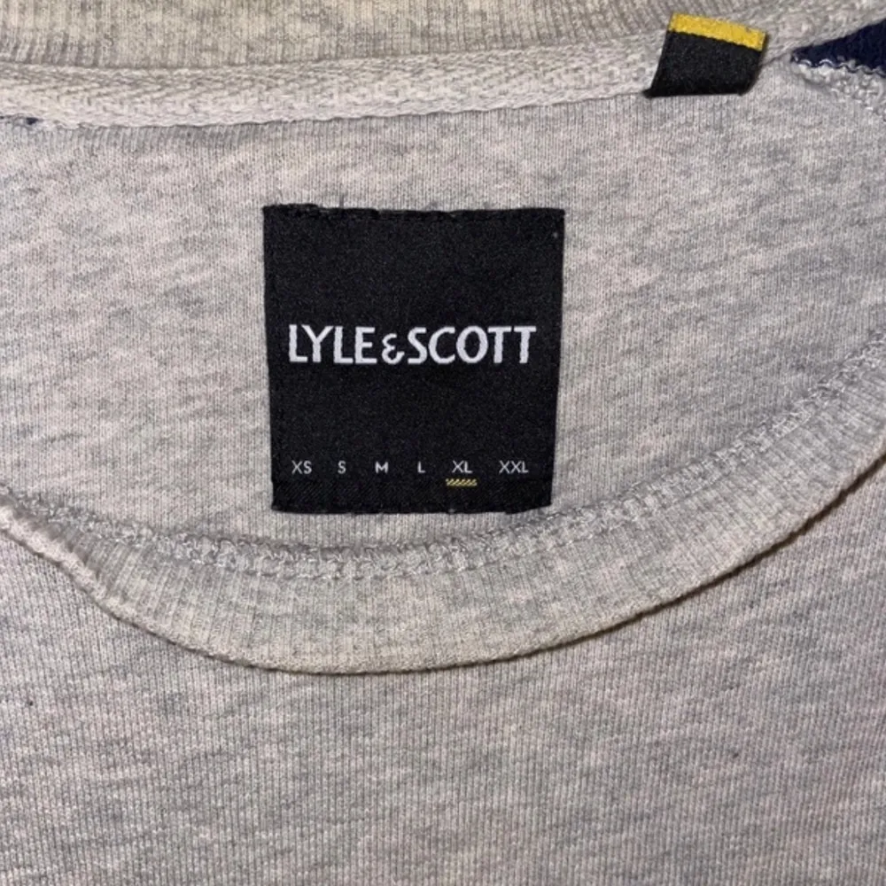 Säljer en snygg grå och marinblå tröja från Lyle & Scott. Den har ett broderat gult emblem på bröstet och långa ärmar. Perfekt för en avslappnad stil under höst och vår. Tröjan är i bra skick och redo för nya äventyr!. Tröjor & Koftor.