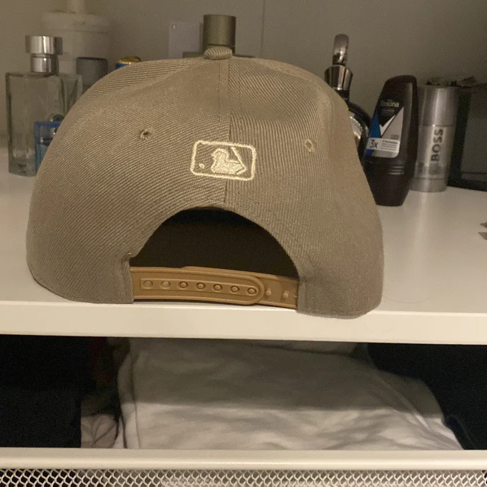 Säljer en beige snapback-keps från New Era med 'A's' broderat framtill. Kepsen har en justerbar rem baktill och MLB-logga. Perfekt för sommardagar eller som en stilren accessoar.. Asusteet.