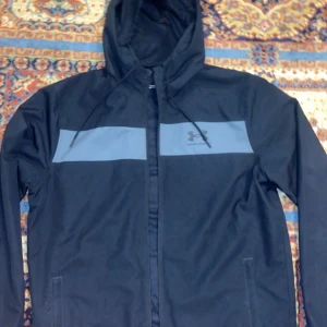 Svart vindjacka från Under Armour - Säljer en svart vindjacka från Under Armour i mycket bra skick. Jackan har en huva och dragkedja framtill, perfekt för höst och vår. Den är tillverkad i syntetmaterial och har en diskret logga på bröstet. Passar både till träning och vardagsbruk.