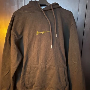 Svart hoodie med gul text från Mennace - Säljer en svart hoodie med en cool gul text på bröstet. Den har en klassisk känguruficka och justerbar huva med snören. Perfekt för en avslappnad stil och passar till det mesta. Skön och bekväm för både vardag och chill.