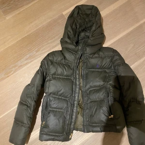 Grön dunjacka från Ralph Lauren - Säljer en snygg grön dunjacka från Ralph Lauren i storlek L i barnstorlek så sitter som en S. Jackan har en praktisk huva och dragkedja framtill. Perfekt för kalla vinterdagar! Den är i mycket bra skick och redo för nya äventyr. 