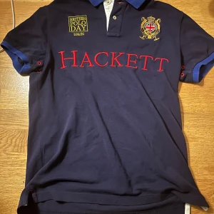 Mörkblå pikétröja från Hackett - Säljer en snygg mörkblå pikétröja från Hackett med röd text och broderad logga på bröstet. Tröjan har korta ärmar med blå detaljer och en klassisk krage. Perfekt för en stilren och sportig look. Passar bra till både vardag och fest! Tröjan är i storlek small och passar bra på en person runt 160-180 cm 