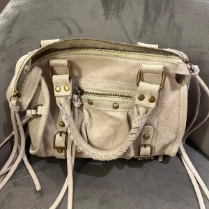 Beige mockaväska med dragkedja - Säljer en snygg beige axelväska i mocka med flera dragkedjor och detaljer i guld. Väskan har flätade handtag och justerbara remmar. Perfekt för både vardag och fest!