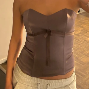 Brun/lila  tubtopp med rosett - Snygg brun tubtopp i ett glansigt silkes material. Toppen har en söt rosett framtill som ger en extra touch. Perfekt för en utekväll eller fest! Passar bra till både jeans och kjol.