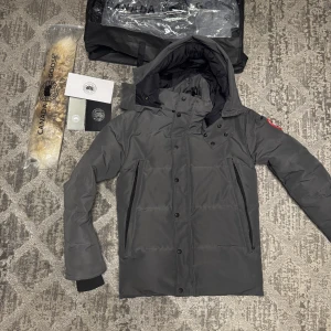 Grå dunjacka från Canada Goose - Säljer en grå dunjacka från Canada Goose i ny skick . Jackan har en avtagbar huva och dragkedja med knappar framtill. Perfekt för kalla vinterdagar. Den har långa ärmar och en klassisk logga på ärmen. Passar både till vardags och mer formella tillfällen. Priset kan diskuteras vid snabba affär