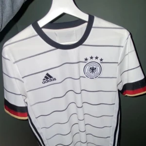 Vit och svart fotbollströja från Adidas - Säljer en snygg vit fotbollströja från Adidas med svarta ränder och Tysklands emblem på bröstet. Tröjan har korta ärmar med ränder i svart, rött och gult. Perfekt för fotbollsfans! 🏆
