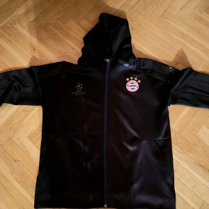 Svart jacka med Bayern München-logga - Säljer en svart Adidas jacka med Bayern München-logga och Champions League-märke. Jackan har en dragkedja framtill och en huva. Den är i bra skick och passar till både vardag och matchdagar.