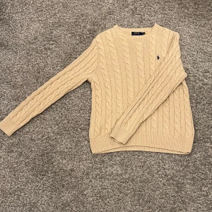 Beige stickad tröja från Ralph Lauren - Säljer en snygg beige stickad tröja från Ralph Lauren och såklart äkta. Den har ett klassiskt flätmönster och den ikoniska loggan broderad på bröstet. Perfekt för höst och vinter. Köpt från butik så kvitto finns, dock får man ej med det då jag köpte fler saker samtidigt. Köpt för 2000kr och aldrig använd då den är lite förstor.