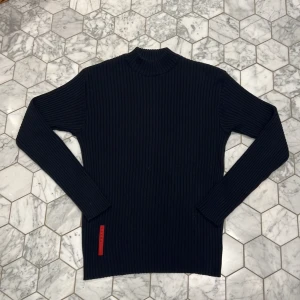 Svart ribbad tröja från Prada - Säljer en stilren svart ribbad tröja från Prada med lång ärm. Perfekt för höst och vinter, den är både varm och bekväm. Tröjan har en diskret röd detalj vid nederkanten som ger en cool kontrast. Passar bra till både vardag och fest!