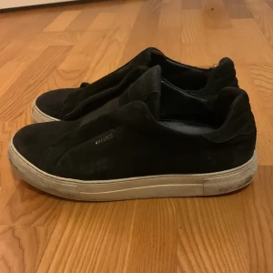 Svarta sneakers från Axel Arigato - Säljer ett par svarta sneakers från Axel Arigato i mocka. Baksidan på ens skorn är lite förstörd men det går att fixa,De har en stilren design med vit sula och är i bra skick. Perfekta för både vardag och fest. Storlek är 42,  men de ser ut att passa en normal passform.