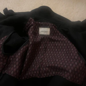 Svart kappa från Ottod'ame - Säljer en elegant svart kappa från Ottod'ame. Den har en klassisk design med knappar framtill och ett bälte i midjan för en snygg passform. Perfekt för höst och vinter, och funkar lika bra till vardags som till fest. Fodret har ett stilrent mönster som ger en extra touch av lyx. Jag är 1.70 och kappan går lite nedanför knäna. En kappa som aldrig går ur tiden. 80 % ull. Nypris ca 3000 kr