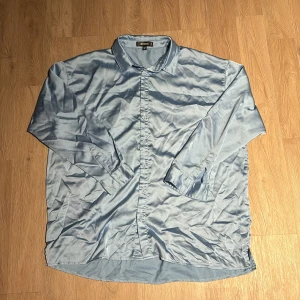 Blå silkesliknande skjorta - Säljer en elegant blå skjorta  i ett silkesliknande material. Skjortan har en klassisk krage och långa ärmar, perfekt för både vardag och fest. Den är i bra skick och ger en stilren look. Passar utmärkt till både jeans och kostymbyxor.