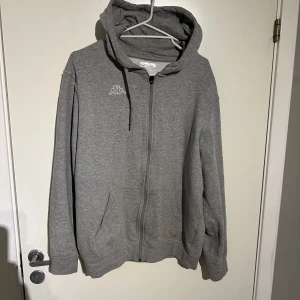 Grå hoodie från Kappa - Säljer en grå hoodie från Kappa i bra skick. Den har en dragkedja framtill och en klassisk känguruficka. Perfekt för höst och vår, med en bekväm passform och långa ärmar. Huvan har dragsko för justering. Perfekt för en avslappnad stil!