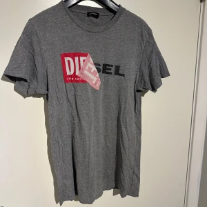Grå t-shirt från Diesel - Säljer en grå t-shirt från Diesel i 100% bomull. Den har en cool logga i rött och svart på framsidan. Perfekt för en avslappnad stil och passar till nästan allt. T-shirten är i nytt skick och har korta ärmar. Perfekt för både vardag och fest!