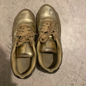 Guldiga sneakers från Nike - Säljer ett par coola guldiga Nike sneakers i bra skick. De har en snygg glansig finish och klassisk snörning. Perfekta för att sticka ut i mängden! Sulan är vit med synlig luftdämpning för extra komfort. Passar både till vardags och fest. Det kan behövas puts.