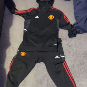 United tracksuit  - Säljer en svart hoodie och matchande byxor från Adidas med Manchester United-logga. Setet har röda detaljer och är perfekt för fotbollsfans. Hoodien har långa ärmar och en bekväm passform. Perfekt för både träning och vardag! Byxorna Storlek M Tröjan S