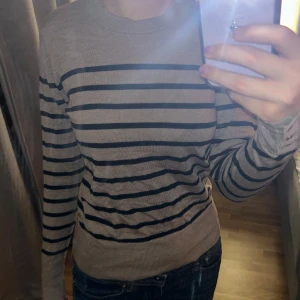 Beige och svart randig tröja - Säljer en snygg beige och svart randig tröja från Lindex! Den har långa ärmar och en normal passform och har även söta gulddetaljer vid slutet av ärmarna. Perfekt för både höst och vår! Tröjan är i bra skick och använd några gånger, inga defekter 💓
