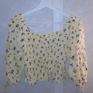 Blommig topp från FB Sister - Söt blommig topp från FB Sister i storlek S. Den är ljusgul med små lila blommor och har en smockad design som ger en fin passform. Toppen har långa ärmar och en lätt volangdetalj vid ärmsluten. Perfekt för vår och sommar!😍