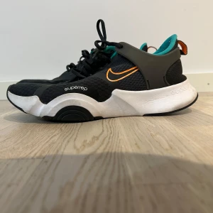 Nike SuperRep träningsskor - Säljer ett par Nike SuperRep träningsskor i svart med orange detaljer. Skorna har en cool design med vit sula och turkos insida. Perfekta för träning med bra stöd och snörning för optimal passform. Använda men i bra skick!