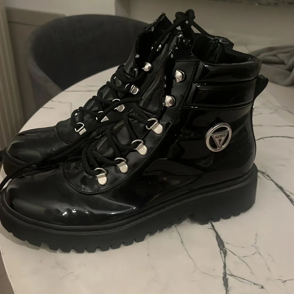 Snygga svarta boots i lackat material från Guess. De har en cool snörning och en praktisk dragkedja på sidan. Perfekta för höst och vinter med en robust sula som ger bra grepp. Styla dem med dina favoritjeans för en trendig look!. Skor.