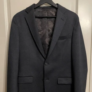 Kavaj från Dressmann stl 48 grå/svart - Säljer denna kavaj från Dressmann i stl 48 (slim fit). Denna väst sitter lika bra på stl M som L. Den har färgen svart/grå och är riktigt lyxig. Kavajen är använd en gång under ett bröllop och är helt ny i skick.