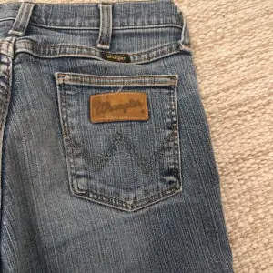 Säljer ett par snygga blå bootcut jeans från Wrangler. De har en klassisk femficksdesign och en rå kant vid bensluten för en trendig look. Perfekta för både vardag och fest! 🕺 säljer dessa för att dom ej kommit till användning då dom är helt oanvända, dom passar mig som är 165. Innerbenslängden är 78cm och midjan rakt över är 34cm