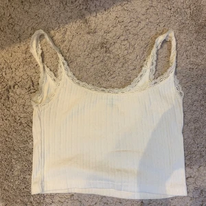 Vit ribbad linnetopp med spetsdetaljer - Säljer en söt vit ribbad linnetopp med spetsdetaljer vid axelbanden och halsringningen. Perfekt för sommardagar eller som ett lager under en kofta. Toppen är i bra skick och har en bekväm passform.