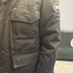 Svart Canada Goose vinterjacka med huva - Säljer en svart vinterjacka i mycket bra skick. Den har en praktisk huva och flera stora fickor framtill. Perfekt för kalla dagar! Jackan har en stilren design med ett märke på ärmen. Passar både till vardags och utomhusaktiviteter.