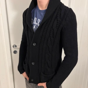 Svart stickad kofta med knappar - Säljer en snygg svart stickad kofta med knappar framtill. Den har ett stilrent flätmönster och en skön krage som ger extra värme. Perfekt för kyliga höst- och vinterdagar. Passar både till vardags och lite finare tillfällen. 🖤