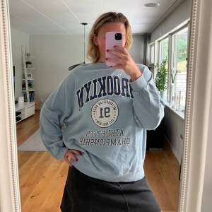 En oversized bekväm ljusblå sweatshirt med texten 'Brooklyn' och 'Athletics Championship' tryckt på framsidan. Tröjan har långa ärmar och ribbstickade muddar vid ärmslut och nederkant. 