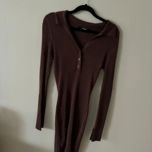 Brun ribbad klänning från BikBok - Säljer en snygg brun ribbad klänning från BikBok i storlek M. Klänningen har långa ärmar och en stilren krage med knappar framtill. Perfekt för höst och vinter, och den är superbekväm att ha på sig. Passar både till vardags och fest!