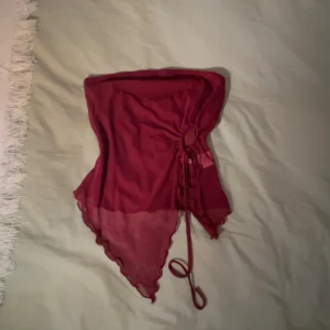Röd axelbandslös topp - Säljer en snygg röd axelbandslös topp med asymmetrisk nederkant och knytning på sidan. Perfekt för festliga tillfällen!
