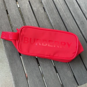 Röd väska från Burberry - Säljer en supercool röd necessär från Burberry. Den är i syntetmaterial och har en dragkedja upptill. är väldigt rymlig går att använda som smink väska eller en vanlig hygiens necessär. Originalpris 450, helt ny och oanvänd