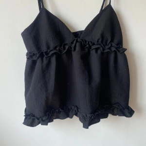 Svart ärmlös topp med volanger - Säljer en superfin svart ärmlös topp med volanger. Den har smala axelband och en lös passform, perfekt för sommardagar eller en festkväll. Materialet känns mjukt och bekvämt.