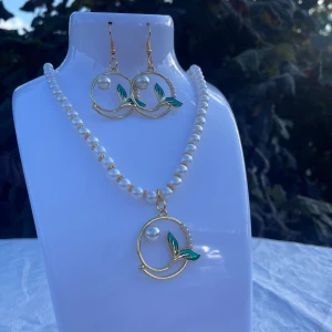 Pärlhalsband och örhängen med gulddetaljer - Säljer ett elegant set med ett pärlhalsband och matchande örhängen. Halsbandet har vita pärlor och ett hängsmycke i guld med en stor pärla och gröna blad. Örhängena har samma design med guldfärgade cirklar, en pärla och gröna blad. Perfekt för en festlig kväll eller som en fin present!
