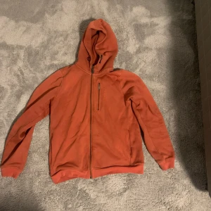 Orange fleecejacka från Sail Racing - Säljer en snygg orange fleecejacka från Sail Racing. Den har en dragkedja framtill och en praktisk bröstficka med dragkedja. Jackan har en huva och långa ärmar, perfekt för kyliga dagar. Den är i storlek 160, vilket passar bra för ungdomar.