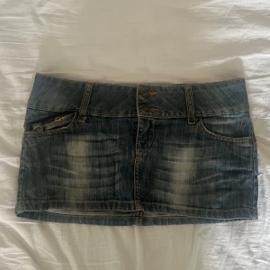 Jeanskjol från SNG Jeans - Säljer en cool låmidjad jeanskjol från SNG Jeans i storlek M. Den är i en klassisk blå denimfärg med slitna detaljer. Kjolen har två knappar fram och dragkedjor både fram och bak. Perfekt för en vanlig dag eller en utekväll! 💗