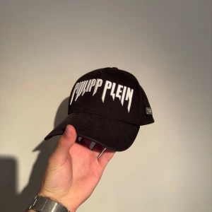 Svart keps från Philipp Plein - Säljer en snygg svart keps från Philipp Plein med vit broderad text på framsidan. Kepsen har en justerbar rem baktill för perfekt passform. Perfekt för att ge din outfit en cool touch!