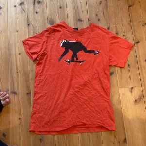 Röd t-shirt med skateboardmotiv från Volcom - Säljer en cool orange/röd t-shirt från Volcom med ett unikt tryck av en apa som åker skateboard. T-shirten är kortärmad och i bomull, perfekt för en avslappnad stil. Volcom-loggan finns tryckt på ärmen. Passar perfekt för skate eller vardagsbruk!