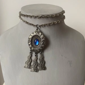 Vintage halsband med blå sten och hängande detaljer - Säljer ett unikt vintage halsband i tenn  med en stor blå sten i mitten. Halsbandet har en vacker kedja och ett hänge med tre hängande detaljer som ger en bohemisk känsla. Perfekt för att ge en extra touch till din outfit!