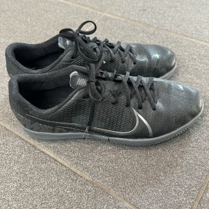 Svarta Nike sneakers - Säljer ett par svarta Nike inomhus skor i bra skick. De har en stilren design med snörning och en grå sula med bra grepp. Perfekta för inomhus träning!