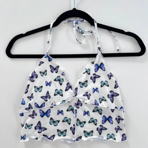 Vit halterneck topp med fjärilar - En vit halterneck topp med fjärilar som knyts runt halsen. Linnet har tunt materiel och är i storlek M 🌟 Säljer det pga att det inte används. Linnet passar mig som normalt har storlek S i toppar.  (ALDRIG ANVÄND)  Längd: 40 cm Byst: 79 cm