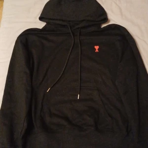 Amiri - Säljer en snygg och bekväm svart hoodie med en liten röd detalj på bröstet. Den har en klassisk känguruficka och justerbar huva med dragsko. Perfekt för både vardag och chill.