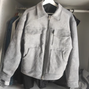 Grå mockajacka med dragkedja - Säljer en snygg grå mockajacka med dragkedja framtill. Jackan har en klassisk krage och praktiska fickor på framsidan. Insidan är fodrad med ett mörkt tyg för extra komfort. Perfekt för höst och vår!
