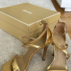Guldiga klackskor från Michael Kors - Säljer ett par snygga guldiga klackskor från Michael Kors. De har en elegant design med smala remmar och en hög klack. Perfekta för festliga tillfällen! Skorna är i storlek 6M och har en glittrig finish som verkligen fångar ögat.