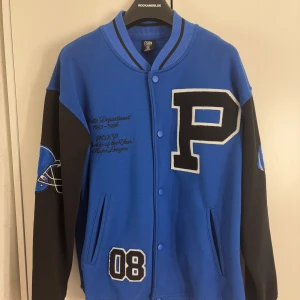 Blå och svart collegejacka från FSBN - Snygg blå och svart collegejacka med stora bokstaven 'P' på bröstet och broderade detaljer. Jackan har långa ärmar och knappar framtill. Den är köpt ifrån Newyorker FSBN, man avdelningen. Men funkat både för kvinnor och män. Den är i storlek XS, och är overzides eftersom det är man storlek. Den har knappar hela vägen och två fickor. 