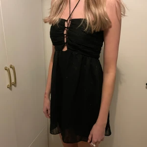 Svart glittrig mini klänning/jumpsuit - Snygg svart kortklänning/jumpsuit med glittrande detaljer och snörning framtill. Klänningen är axelbandslös och har en elegant passform som passar perfekt för festliga tillfällen.