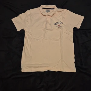 Beige pikétröja med Cape Cod-tryck - Snygg beige pikétröja med korta ärmar och klassisk krage. Tröjan har ett stilrent Cape Cod-tryck på bröstet. Perfekt för en avslappnad stil.