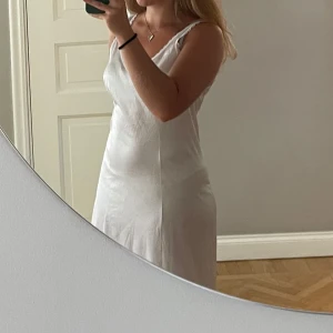 Vit långklänning med spetsdetaljer - Elegant vit långklänning med v-ringning och spetsdetaljer längs axelbanden. Klänningen har en smickrande passform och är perfekt för speciella tillfällen.